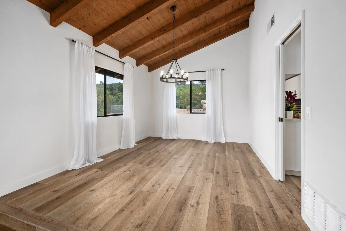 Empty room with wooden floor, white walls, white curtains, windows showing greenery outside, and wooden ceiling beams with a chandelier.