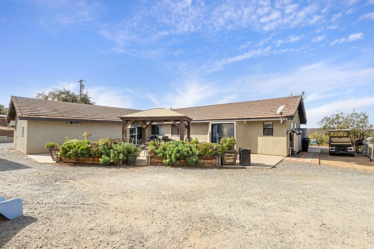 Backyard of a house with a gravel ground, a covered patio area with outdoor furniture, and garden beds with green plants. The house has beige stucco walls and a tiled roof, with a blue sky overhead.