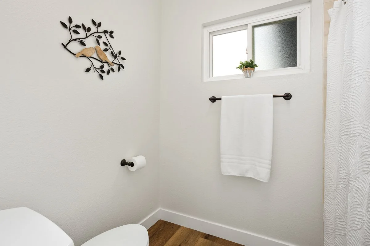 Bathroom with small window, white walls, black towel rack with a white towel, black toilet paper holder with tissue, decorative wall art with two birds and leaves, small potted plant on window ledge, white shower curtain, wood floor