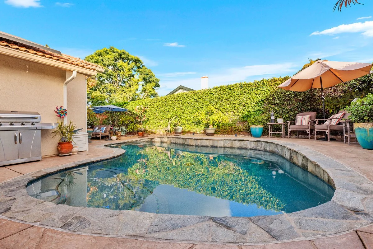 Backyard with a kidney-shaped swimming pool, surrounded by potted plants, lounge chairs, tables with umbrellas, and a stone patio. There is a hedge in the background and a clear blue sky.