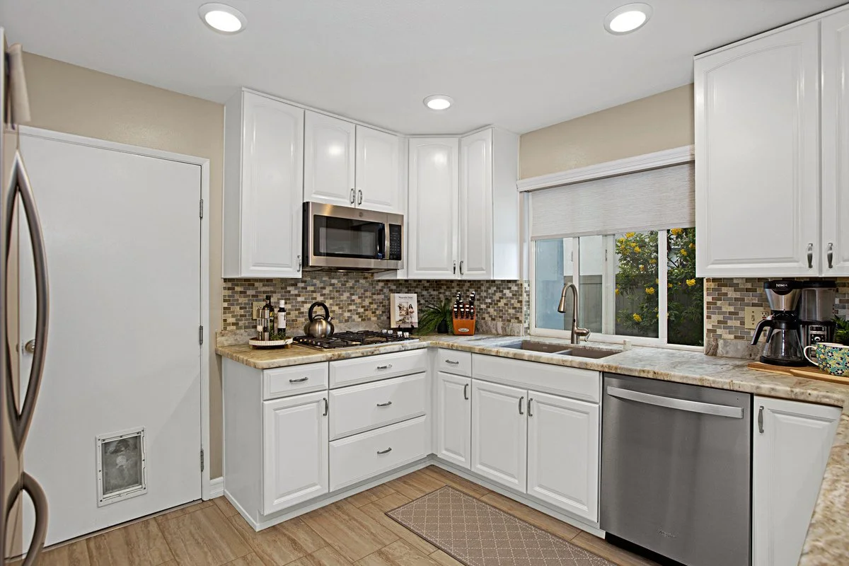 Kitchen with white cabinets, beige countertops, a mosaic tile backsplash, a stainless steel microwave, dishwasher, faucet, coffee maker, and a window overlooking a garden.
