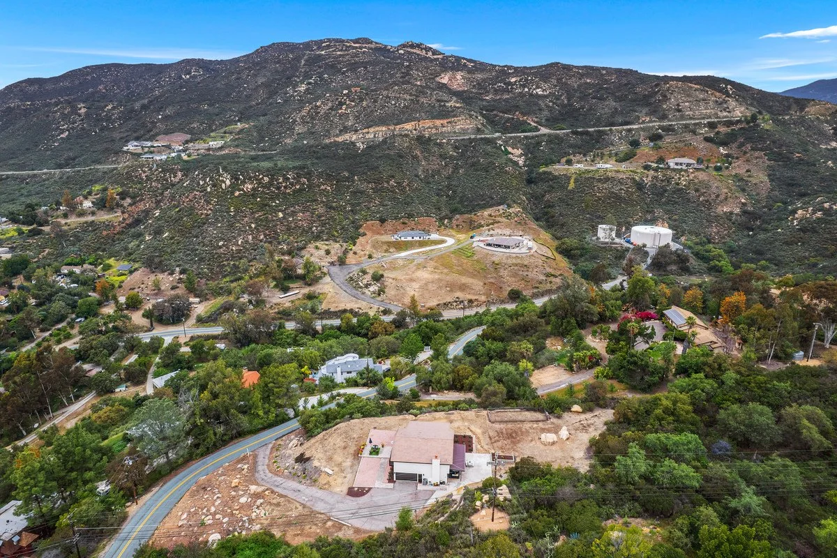 Aerial view of a hilly residential area with winding roads, houses, and green trees against a mountain backdrop.