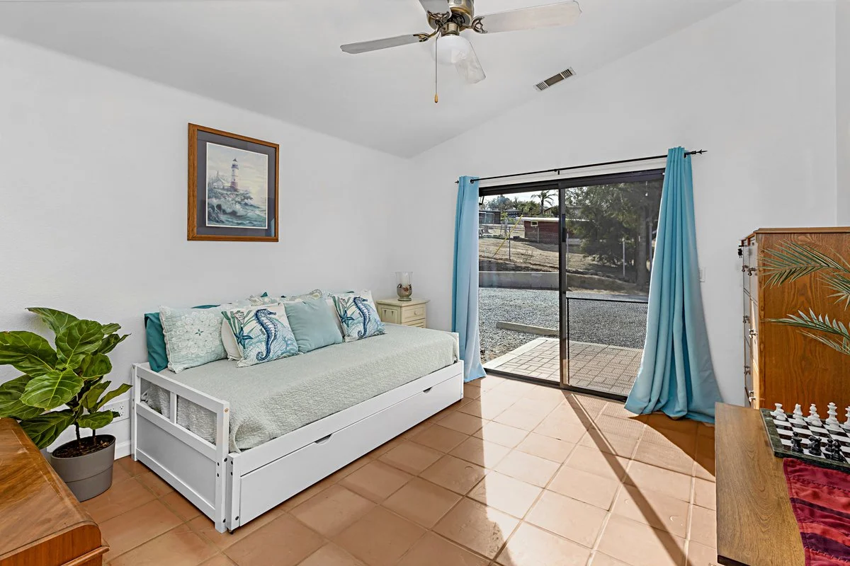 A bright, white-walled living room with terracotta tile flooring. There is a white daybed with ocean-themed pillows, a potted plant on a wooden table, and a small beige nightstand with a glass vessel on top. Blue curtains frame a sliding glass door t