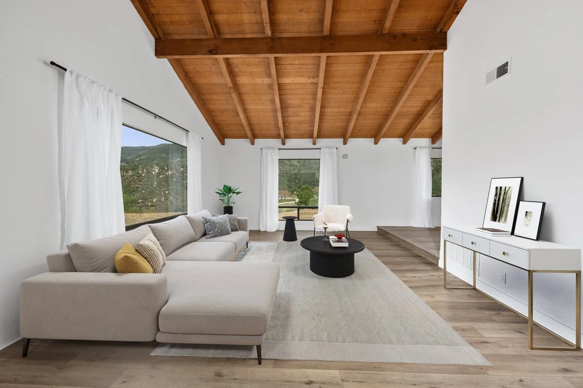 Bright living room with white walls, large windows with white curtains, a beige sectional sofa with pillows, a white armchair, a black round coffee table, a light-colored rug, a white sideboard with framed pictures, potted plant, and a wooden ceiling