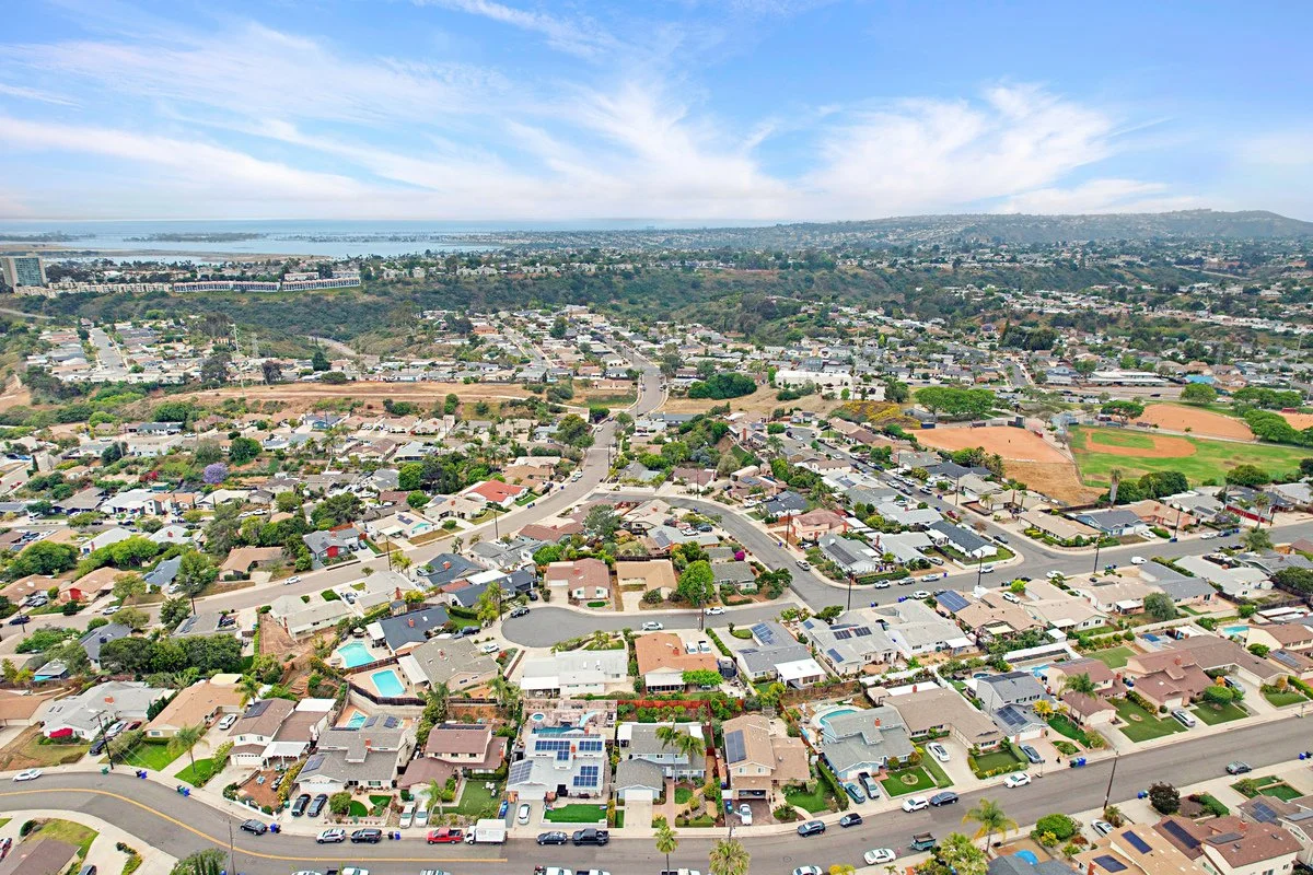 Aerial view of a suburban neighborhood with houses, streets, trees, a swimming pool, and nearby parks, with a body of water in the background.
