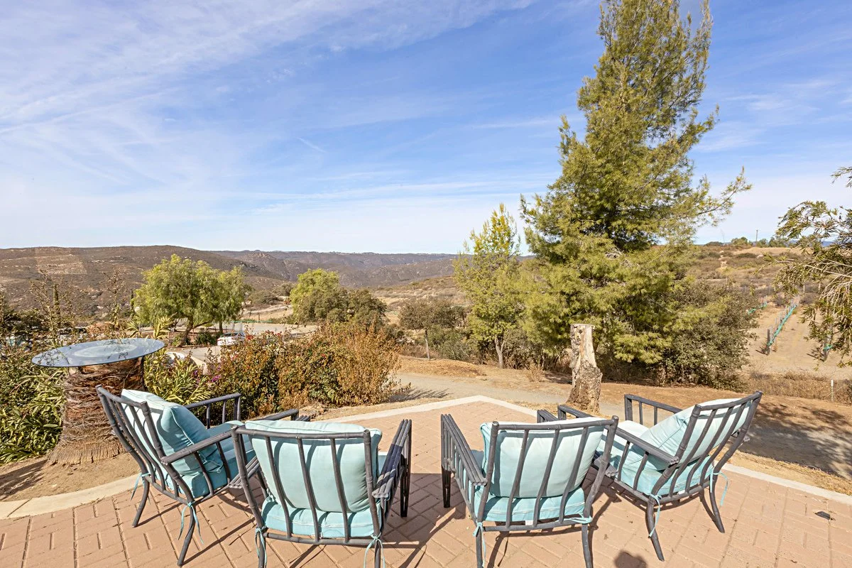 Patio with four black metal chairs and light blue cushions, glass side table, dry landscape with trees, shrubs, and hills in the distance under a blue sky.