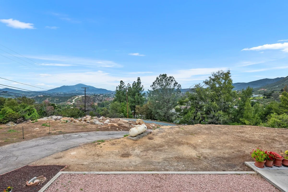 View of a backyard with a dirt patch, some potted plants, and a propane tank, overlooking a hilly landscape with trees and mountains under a blue sky with clouds.