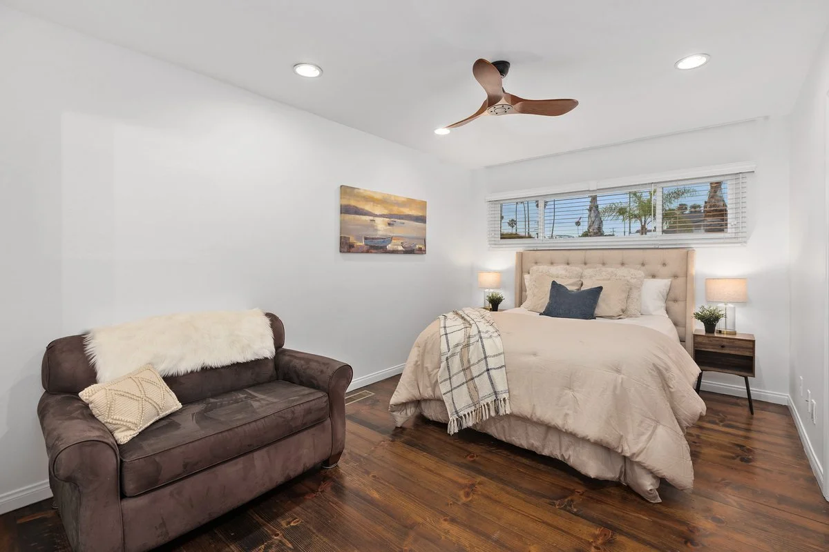 A cozy bedroom with a beige bed, navy blue pillows, white walls, and a window showing palm trees outside. There's a brown sofa with a white furry throw and a cream pillow. Nightstands with lamps and plants are on each side of the bed, and a ceiling f