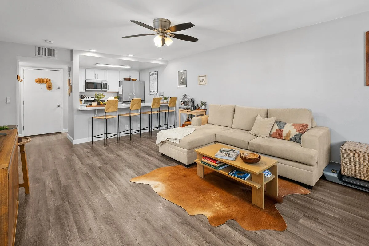 A living room with a beige sectional sofa, wooden coffee table, cowhide rug, and a ceiling fan, with an open kitchen in the background featuring a breakfast bar with four yellow chairs.