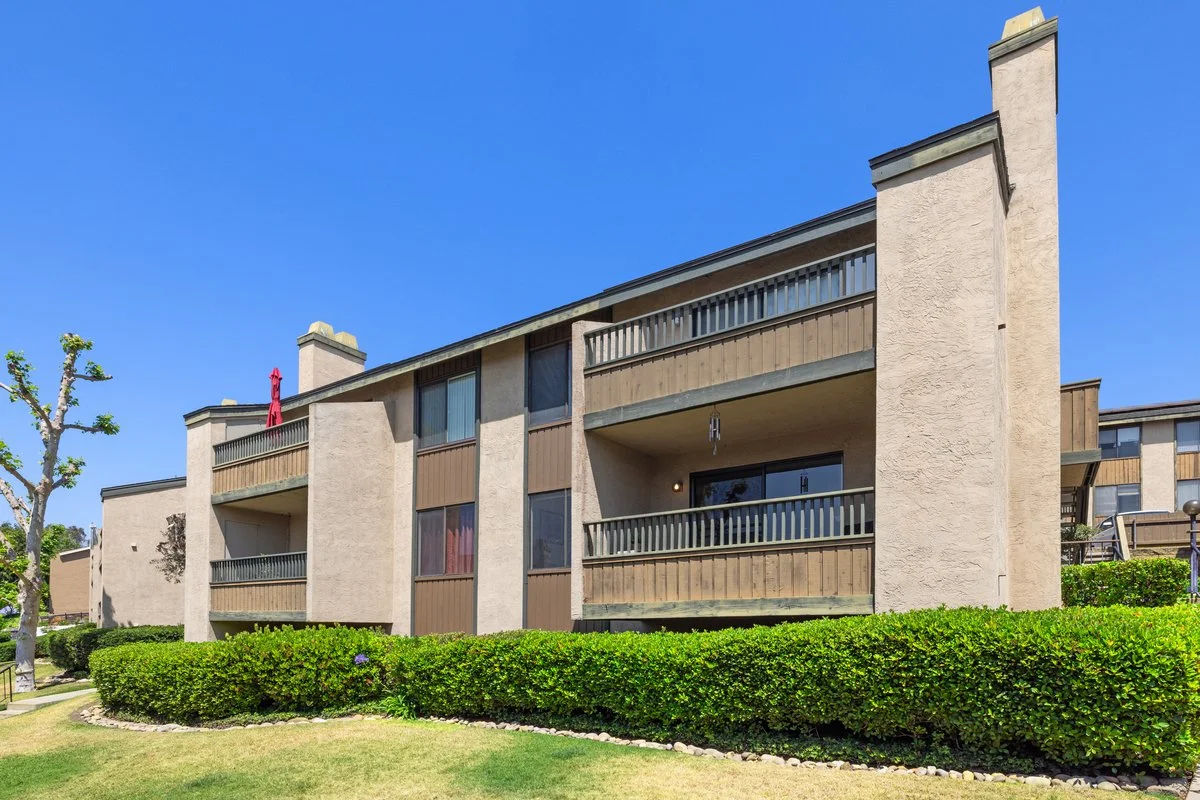 Exterior of a multi-story residential apartment building with balconies, a blue sky, and well-maintained landscaping.