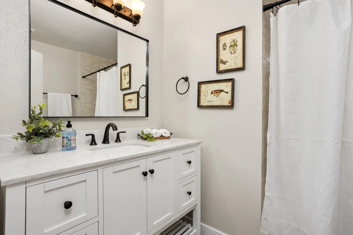 A bathroom vanity with a white cabinet, a marble countertop, a black faucet, a mirror, a framed artwork on the wall, a plant, a soap dispenser, and a towel on a rack.