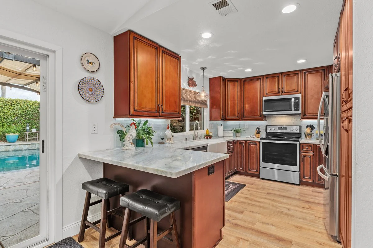 Kitchen with wooden cabinets, stainless steel appliances, marble countertop bar with two black stools, and a view of a backyard with a pool through a glass door.