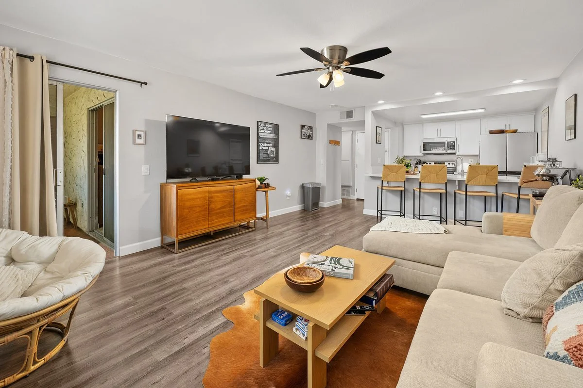 Living room with a beige sofa, armchair, wooden coffee table, large flat-screen TV on a wooden stand, ceiling fan, open kitchen with barstools, and sliding glass door with curtains.
