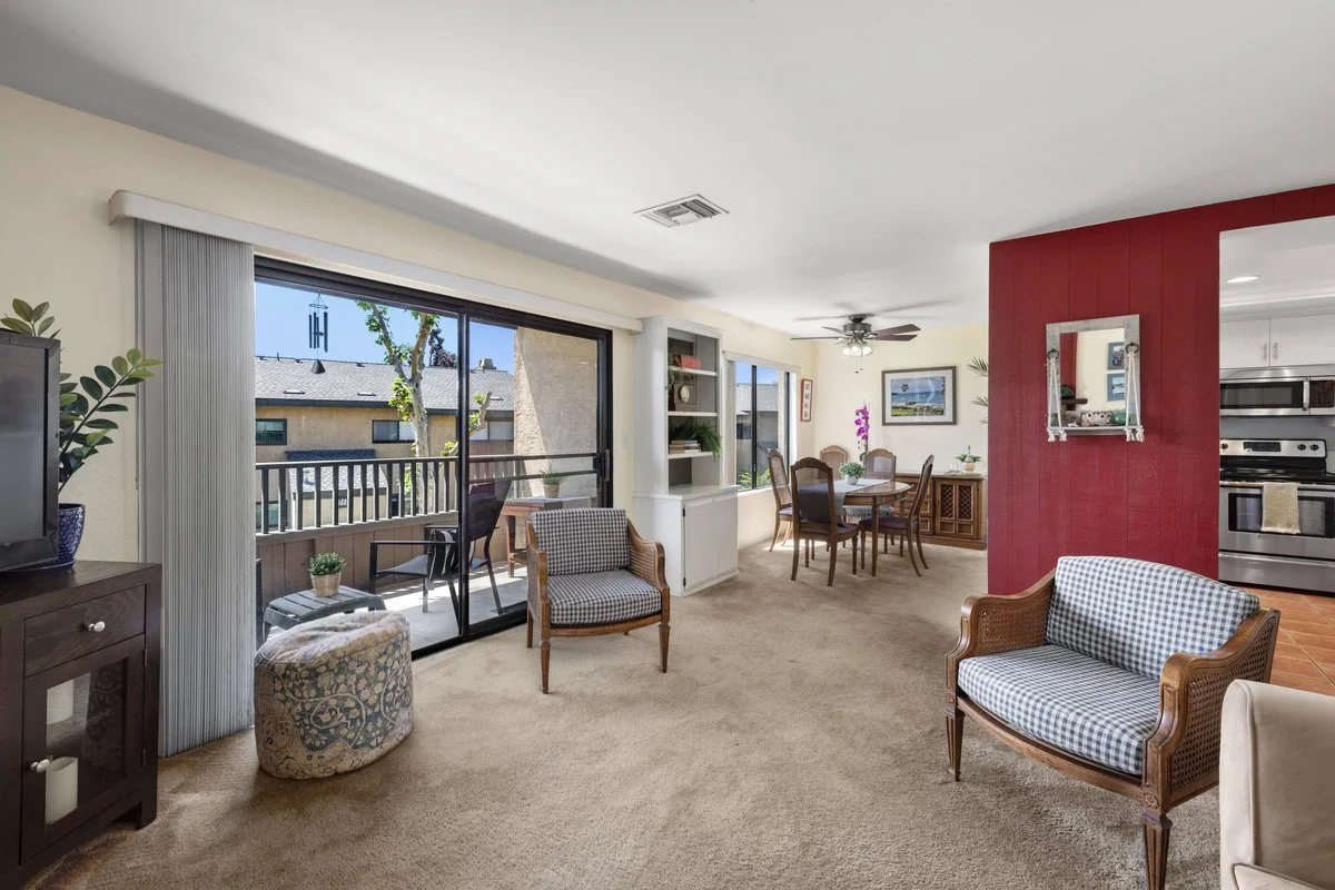Living room with sliding glass door leading to balcony, dining area, and kitchen, featuring cushioned chairs, a red accent wall, and decorative plants.