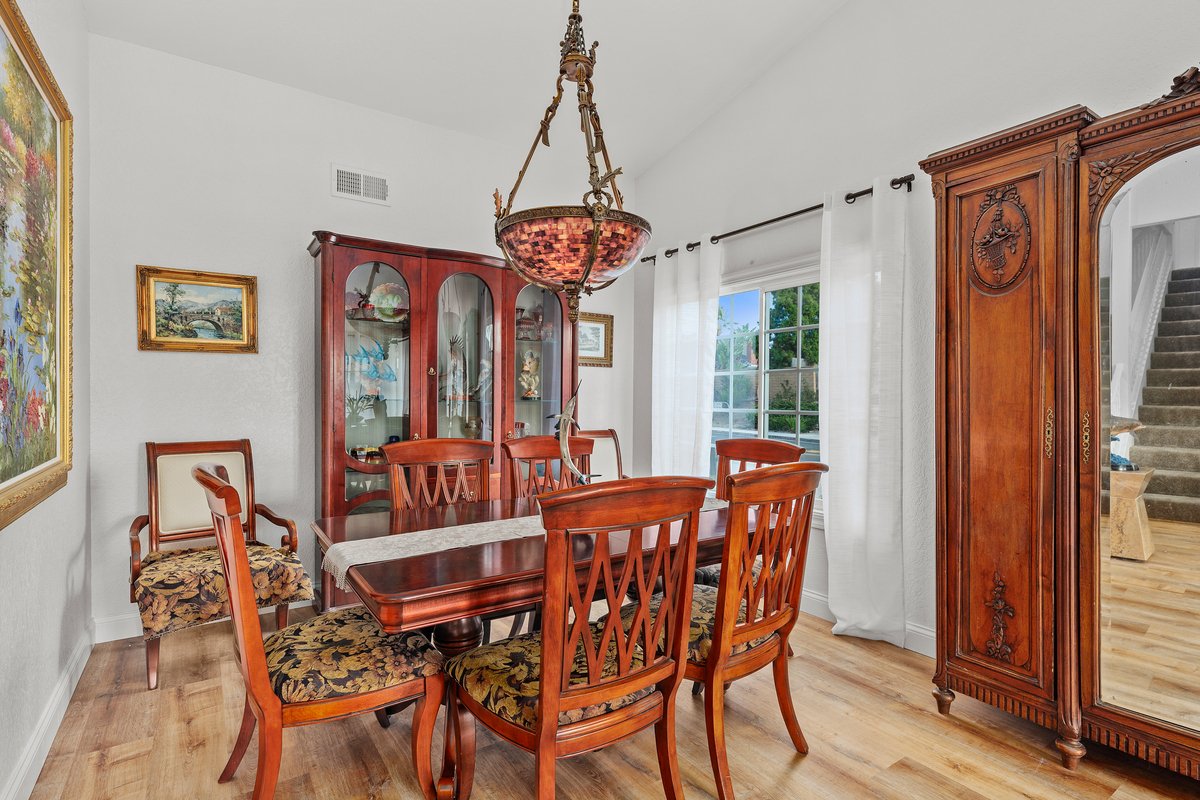 A dining room with a wooden table and six matching chairs with patterned cushioned seats. A glass-front china cabinet, an ornate wooden armoire, a window with white curtains, and a chandelier hanging above the table.