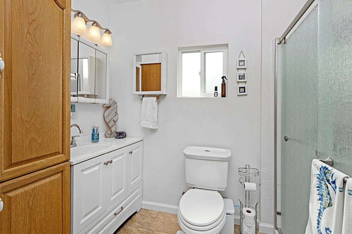Bathroom with white walls, wood cabinets, a small mirror, a window, a toilet, a shower with frosted glass door, and hand towels hanging on a towel bar.