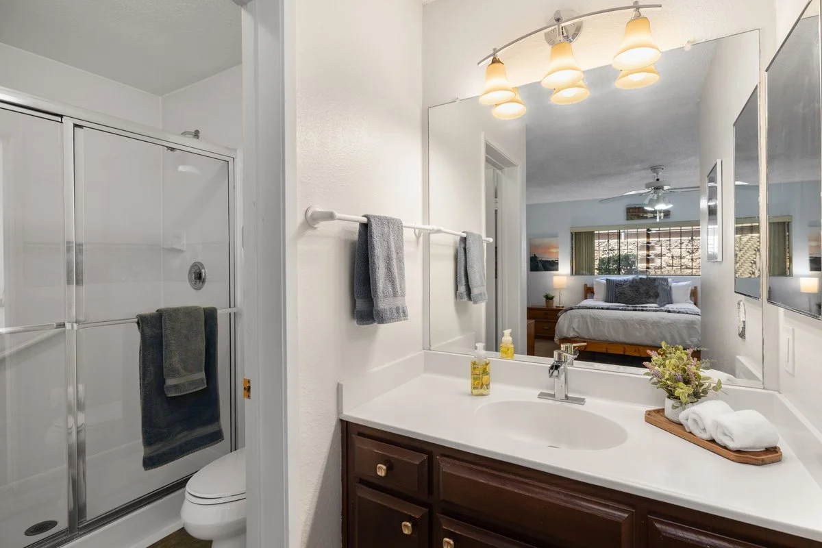 Bathroom with a mirror, sink, and vanity. The room reflected in the mirror shows a bedroom with a bed, nightstands, lamps, and a large window.