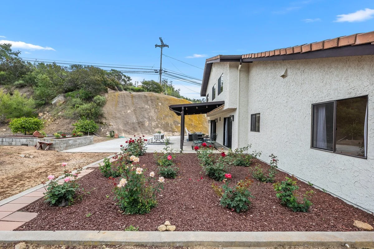 Backyard of a house with rose bushes and a patio area with outdoor furniture, hillside with power lines in the background.