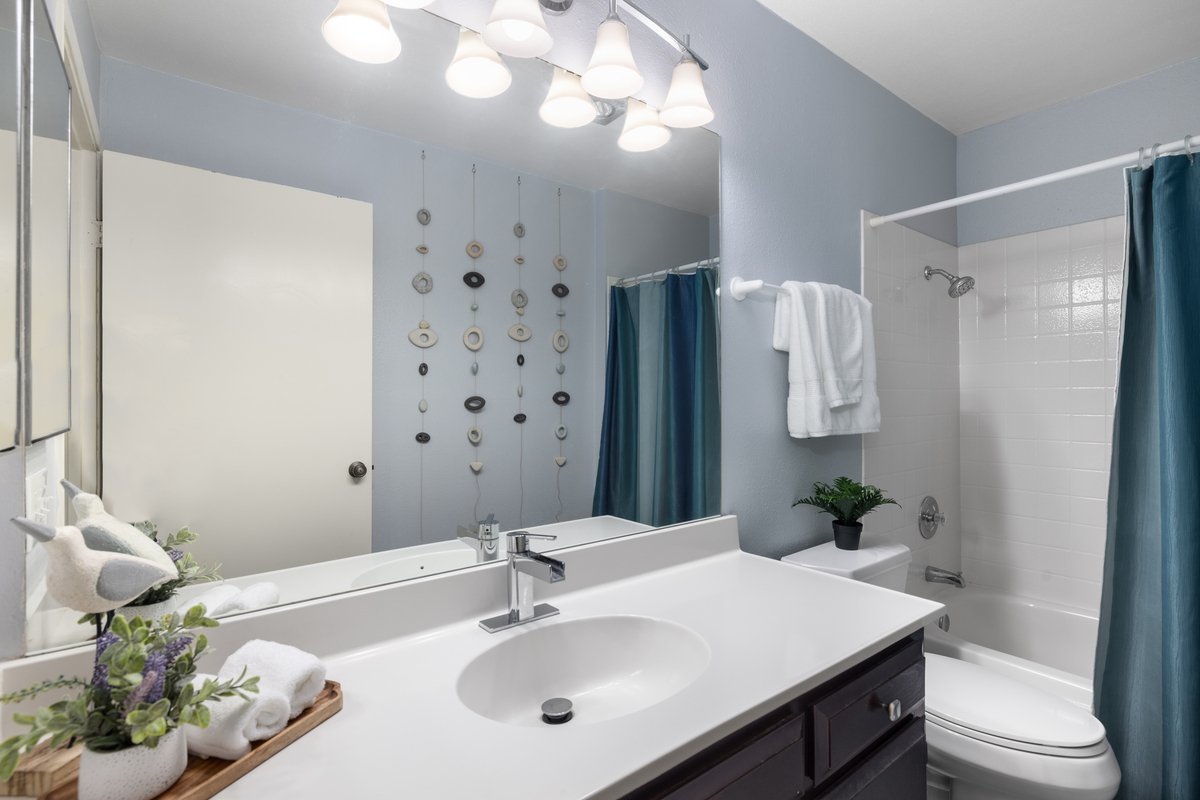 Bathroom with a white vanity, mirror, shower with a blue curtain, a wall decoration, and a towel hanging, with a plant and a plush bird decoration on the counter.