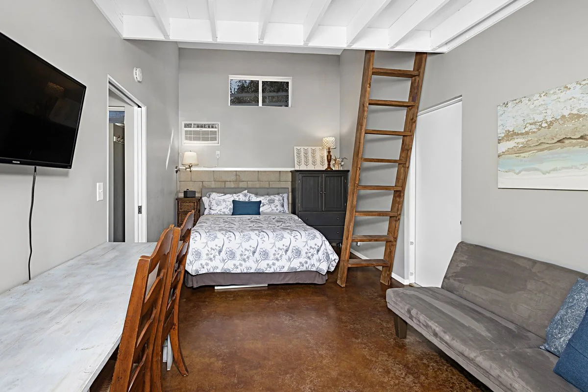 A cozy studio apartment with a bed, desk, TV, ladder, and small living area.