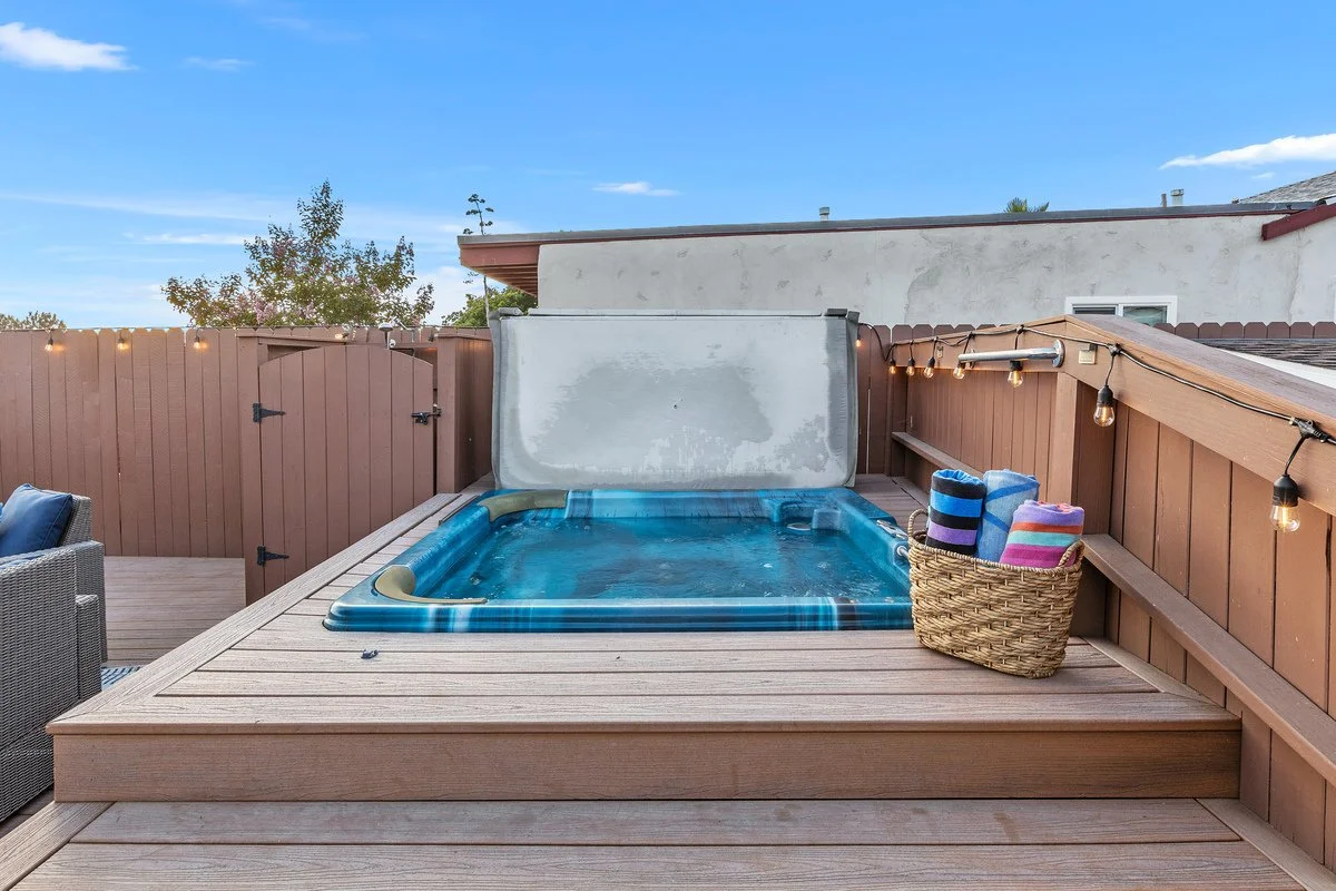 A backyard deck with a hot tub partially covered by a lid, surrounded by brown wooden fencing and string lights, with a basket of colorful towels placed on the deck.