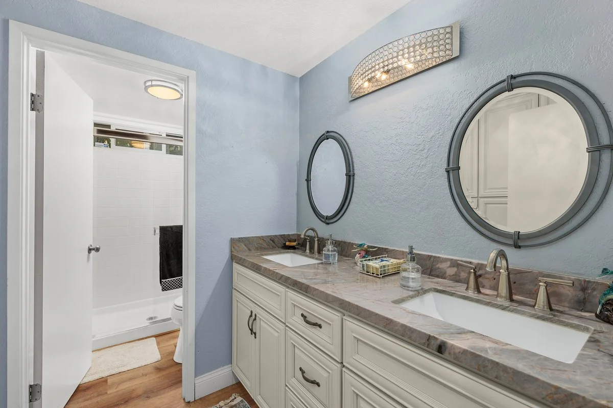 Bathroom double sink vanity with oval mirrors, blue textured wall, and a tiled countertop, adjacent to a walk-in shower with white tiled walls.