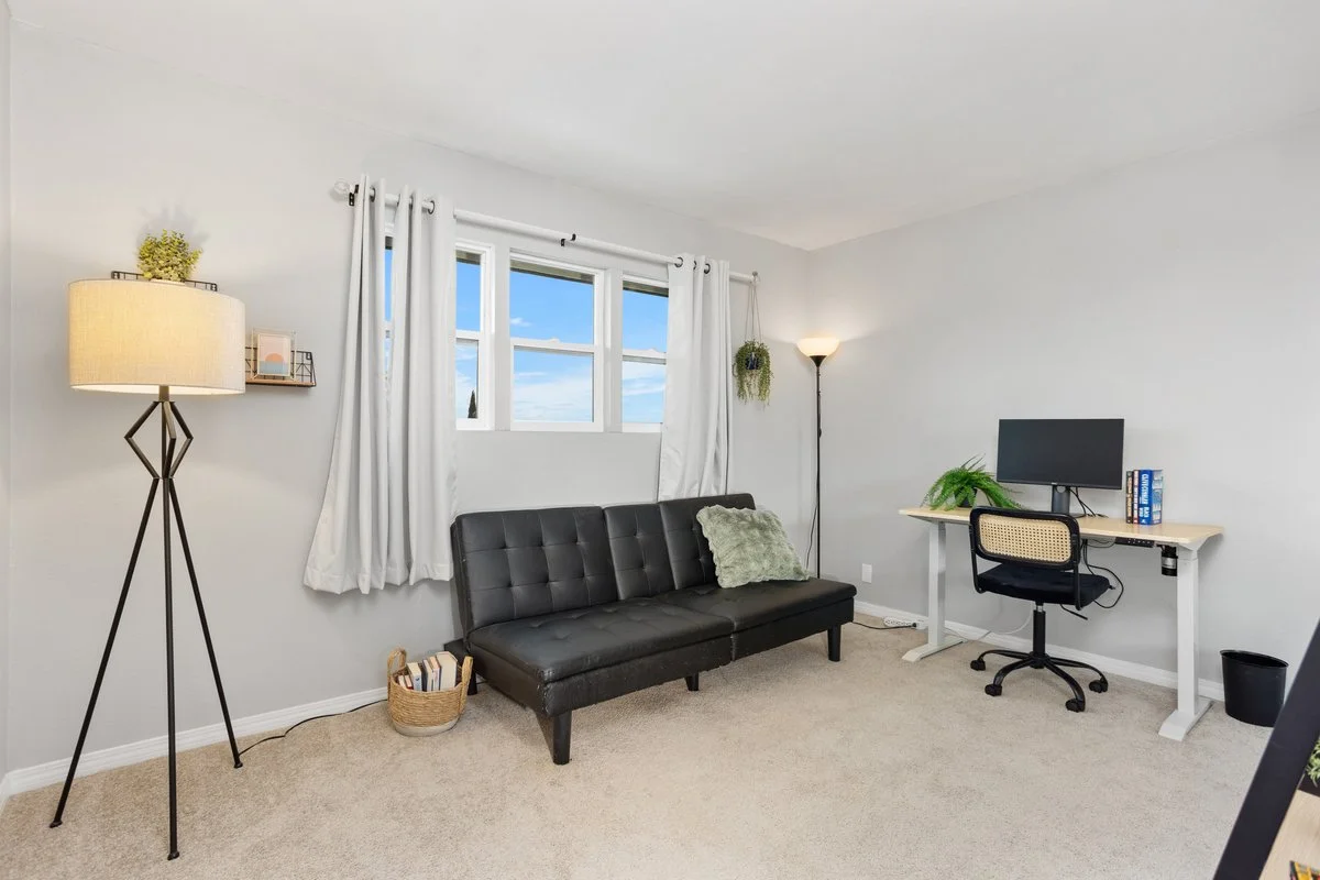 A bright, minimalist living room with white walls, a window with white curtains, a black futon sofa with a green pillow, a tall floor lamp, a small desk with a computer monitor, and a black swivel chair.