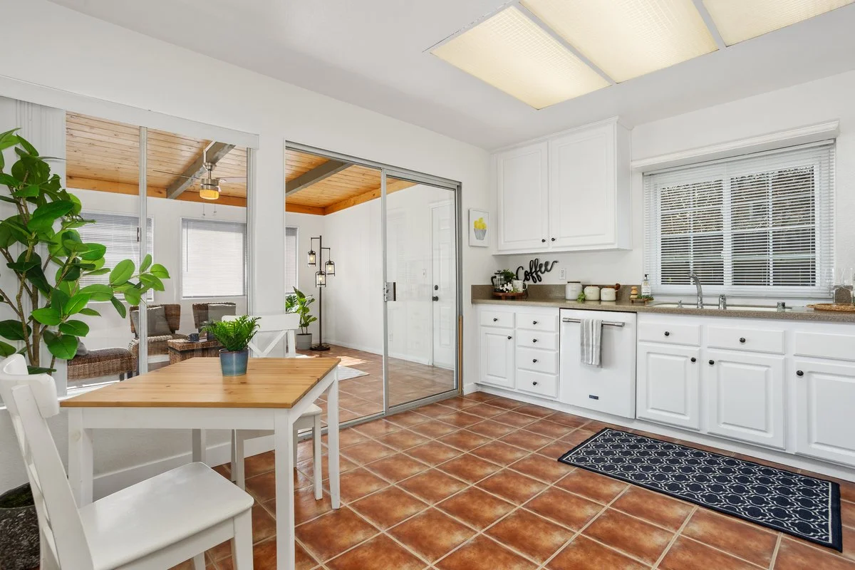Kitchen with white cabinets, brown countertops, ceramic tile flooring, and blinds on the windows. A small dining table with a plant in a pot, and a glass sliding door leading to a sunroom with wicker chairs. Ceiling lights and a small decorative sign