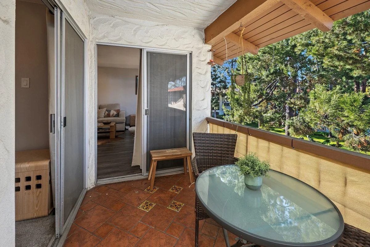 Balcony with a glass-top table with a potted plant, a wicker chair, hanging plant, and view of green trees outside.