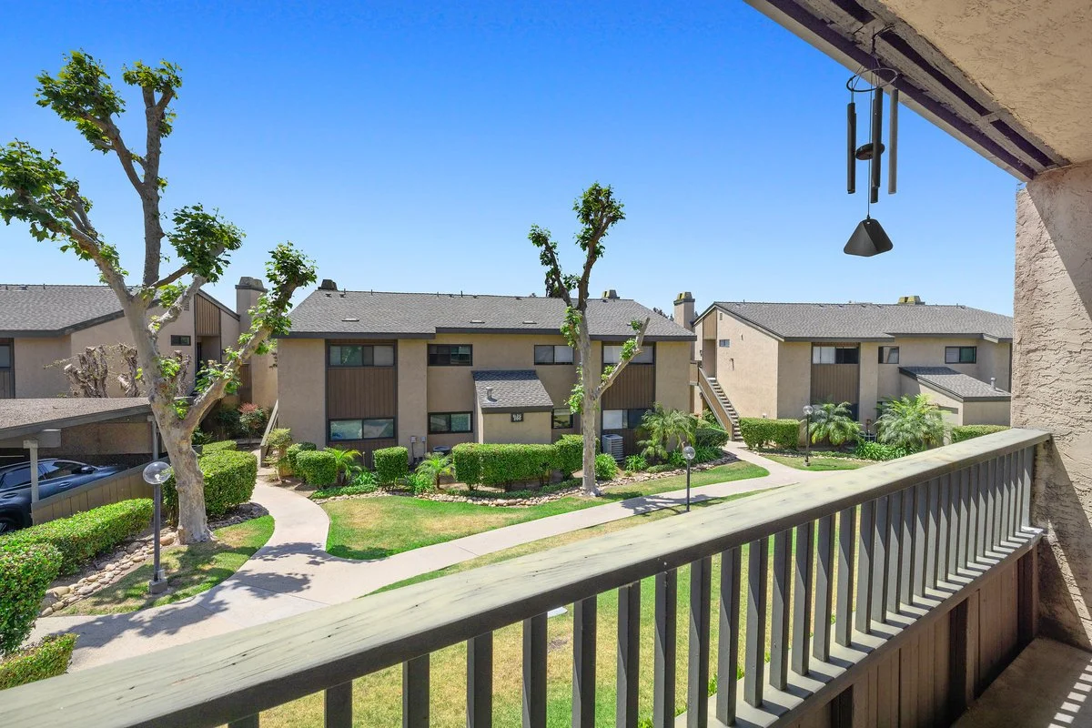 View from a balcony overlooking a residential apartment complex with landscaped gardens, pathways, trees, and multiple buildings under a clear blue sky.