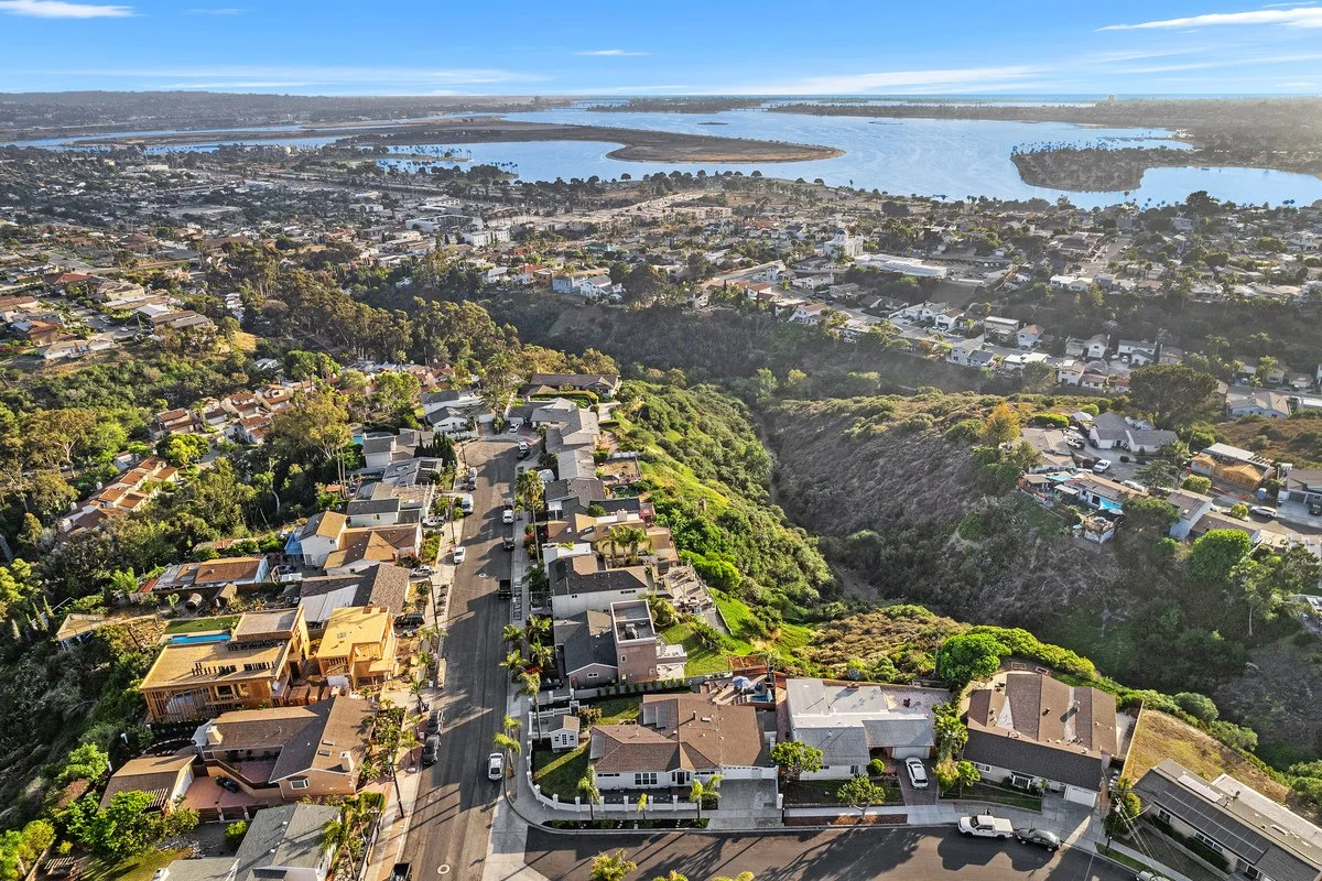 Aerial view of a residential neighborhood with houses, trees, and a street, overlooking a body of water with islands and land formations in the distance.