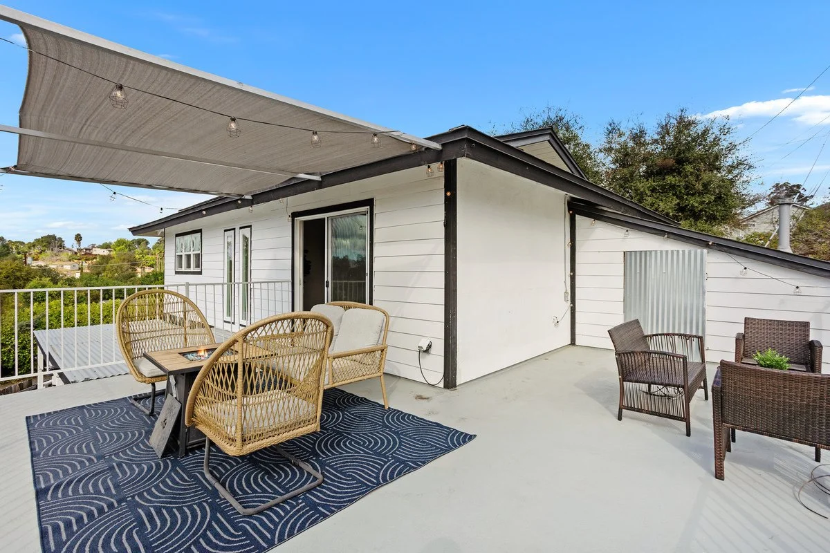 Outdoor patio with wicker and metal chairs, a small table, a blue patterned rug, string lights overhead, and a white house with black trim in the background.