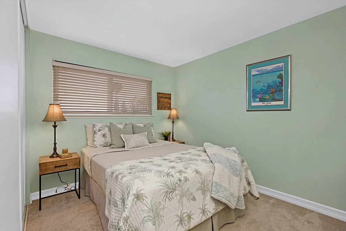 A cozy bedroom with a bed, two wooden nightstands, and lamps. The bed has palm tree patterned bedding and pillows. There is a window with closed blinds and a framed picture on the wall.