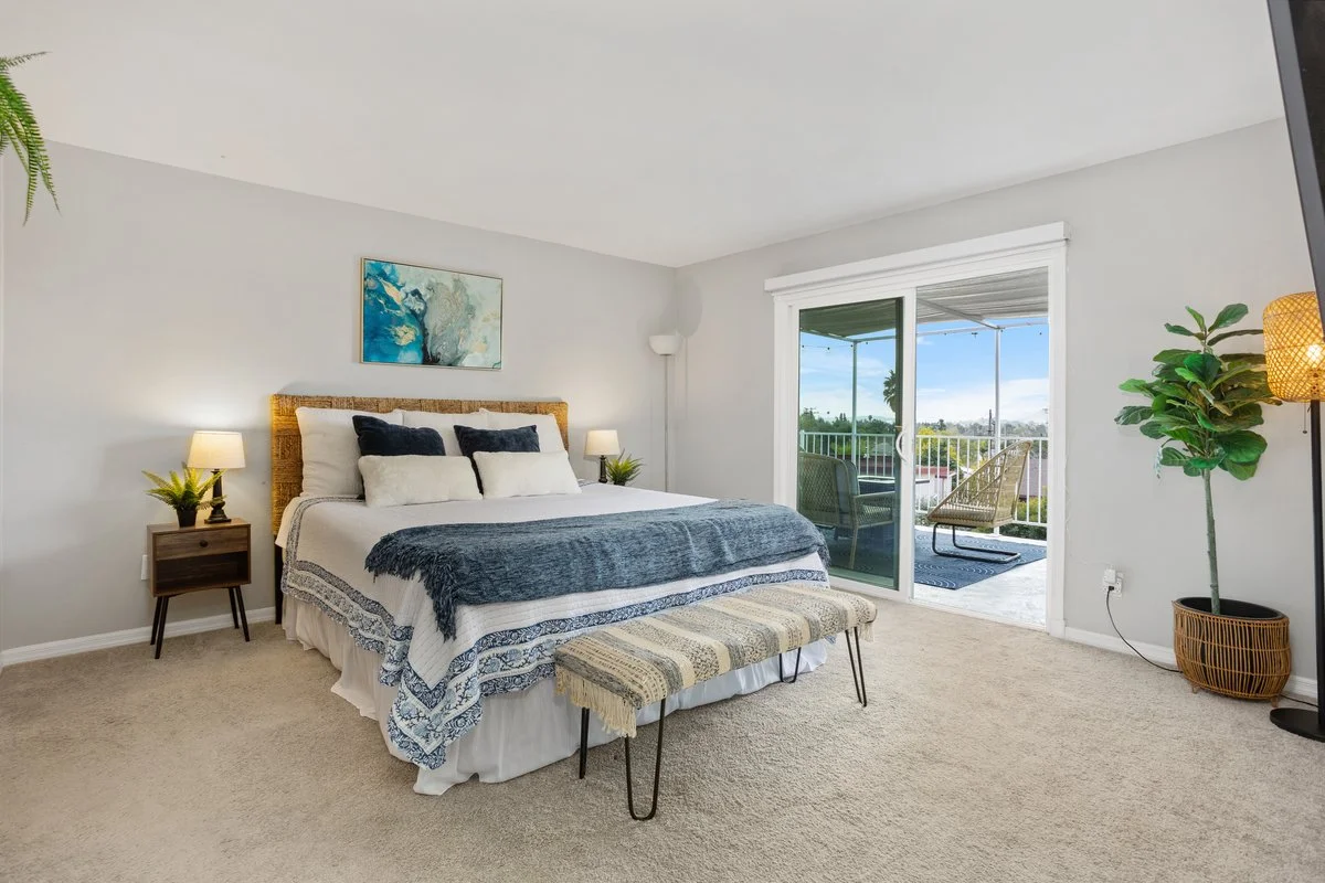 A bedroom with a large bed, nightstands with lamps, a bench at the foot of the bed, and a sliding glass door leading to a balcony with outdoor seating and a view of trees and sky.