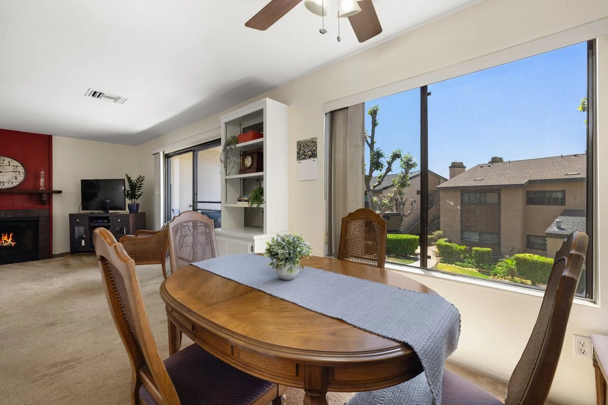 Living room with large window, dining table with wood finish, white vase with greenery, view of residential neighborhood outside, television, fireplace, ceiling fan.