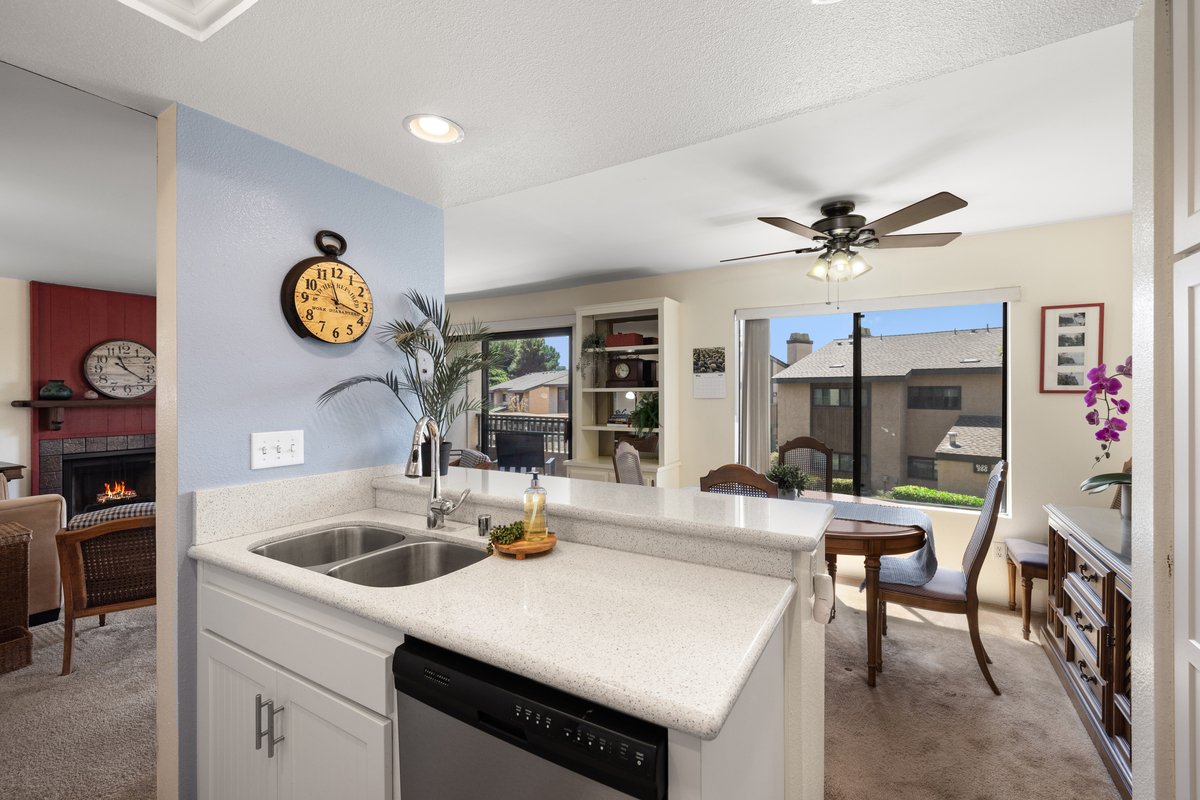 Open-concept living area with white kitchen island, dining table with chairs, and a living room with a fireplace, large windows, and ceiling fan.
