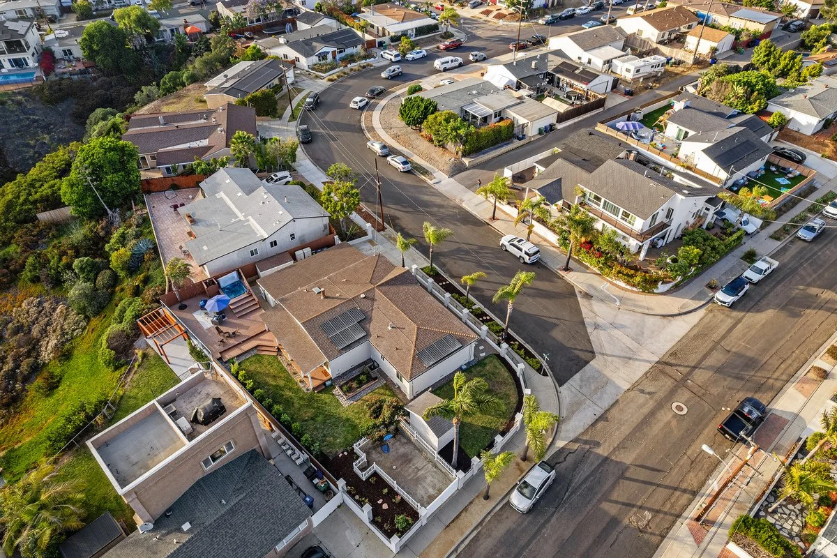 Aerial view of a suburban neighborhood with single-family homes, streets, parked cars, and lush trees.