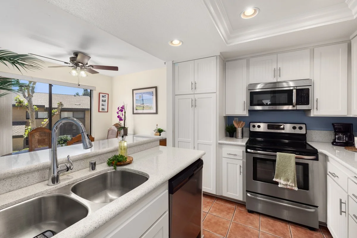 Kitchen with white cabinets, stainless steel stove and microwave, black coffee maker, white countertops, terracotta tile floor, and a view into the dining area with a sliding glass door and outdoor seating.