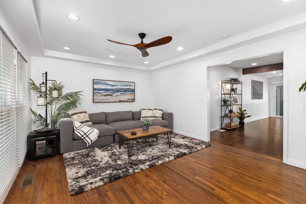 Living room with a gray sofa, a wooden coffee table, a patterned rug, a tall black lamp with plants, a framed landscape painting, white walls, and hardwood floors.