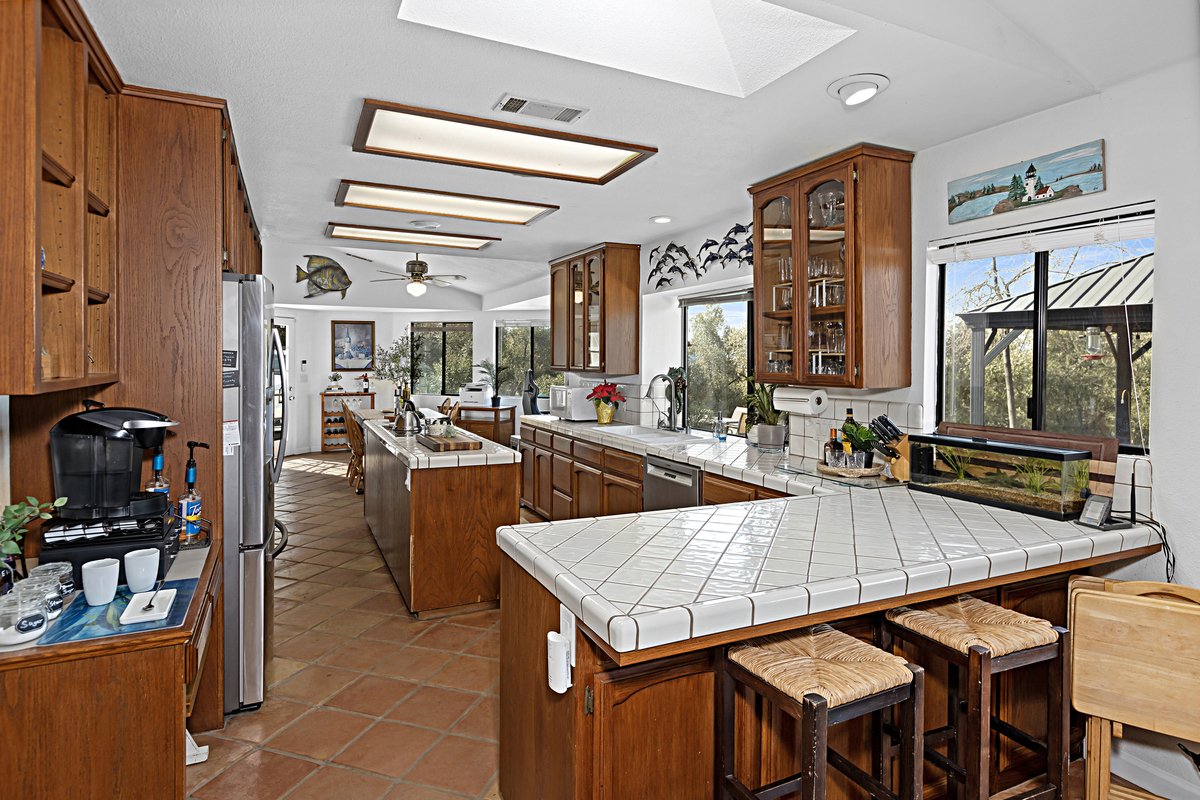 A spacious kitchen with wooden cabinets, white tiled countertops, a stainless steel refrigerator, a fish decoration on the wall, and large windows showing trees outside.