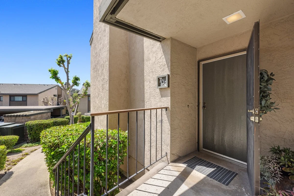 Apartment entrance with a screened door, house number 70, and a small outdoor light. A concrete walkway leads to the door, with greenery and bushes nearby, and neighboring apartment buildings in the background under a clear blue sky.