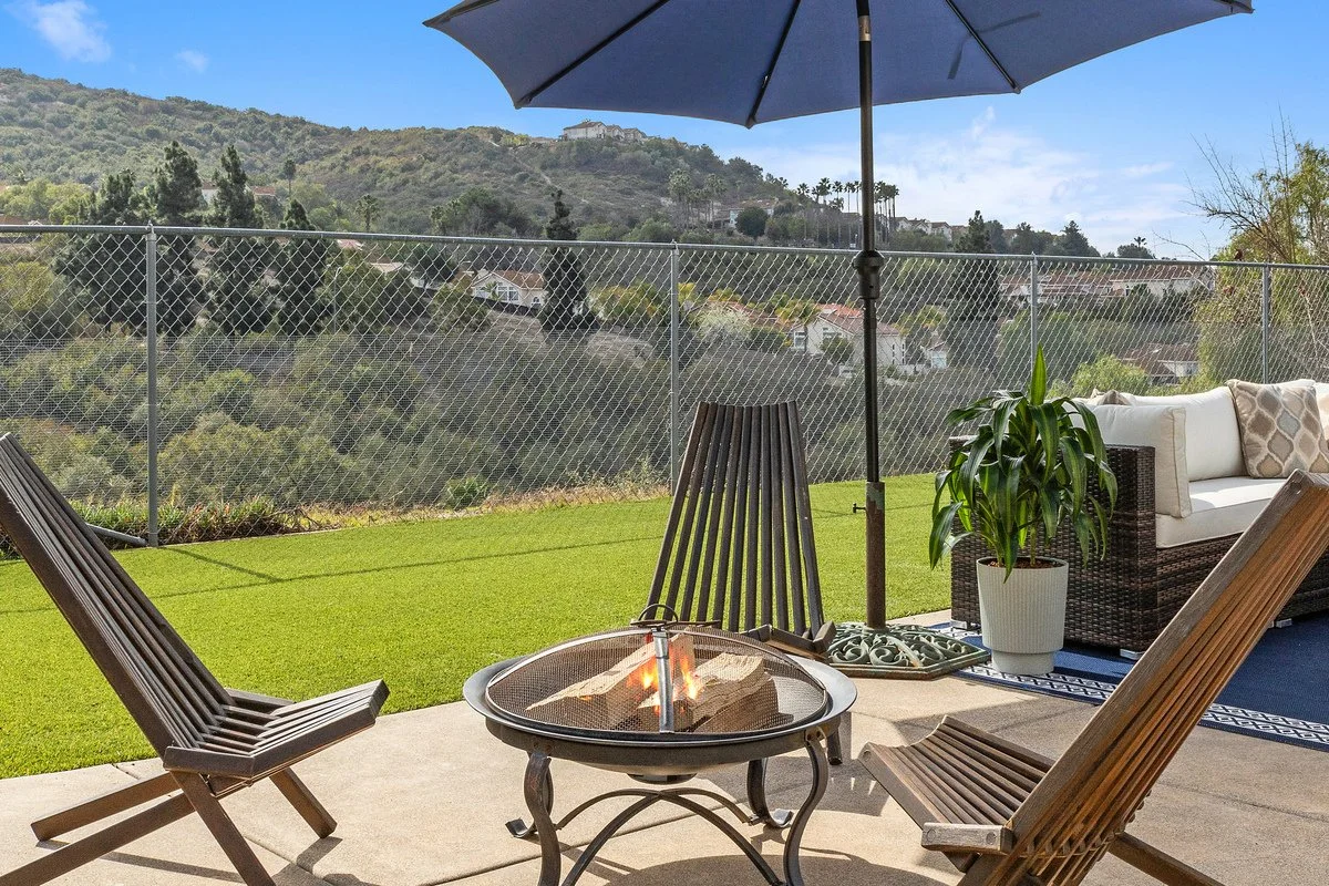 Outdoor patio with a fire pit, wooden chairs, a large patio umbrella, a potted plant, a wicker couch with cushions, on a sunny day with hills and houses in the background.