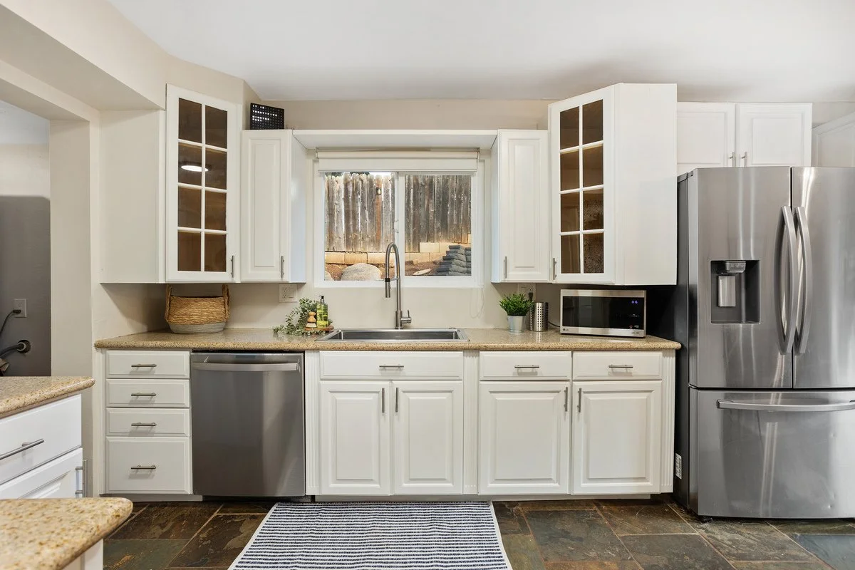 Kitchen with white cabinets, stainless steel refrigerator, microwave, and dishwasher. There is a window with a view of a wooden fence and outdoor area, with various plants and small stones outside.