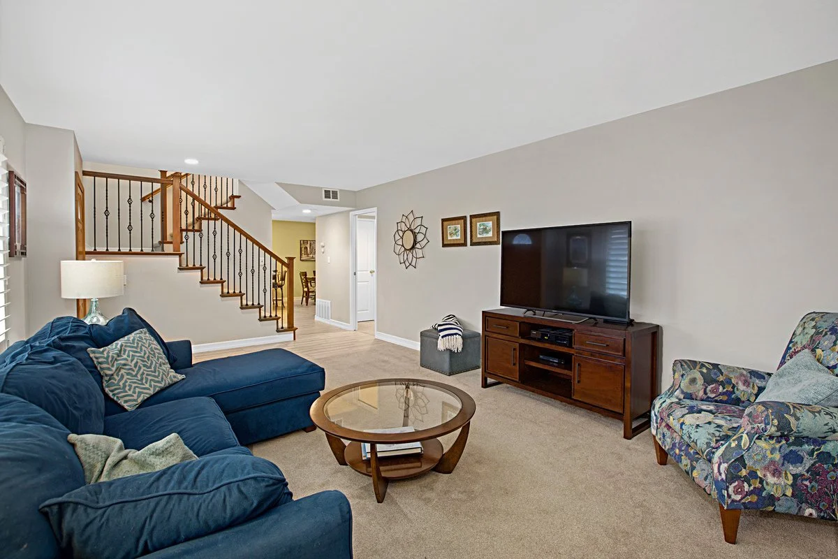 Living room with blue sofa, patterned armchair, wooden coffee table, TV on wooden stand, wall decorations, lamp, stairs, and carpeted floor.