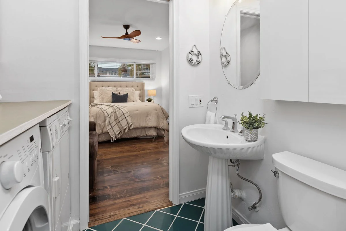Bathroom with white walls, a pedestal sink, a mirror, a toilet, and a small potted plant. Visible laundry appliances on the left, bedroom with a bed and window in the background.