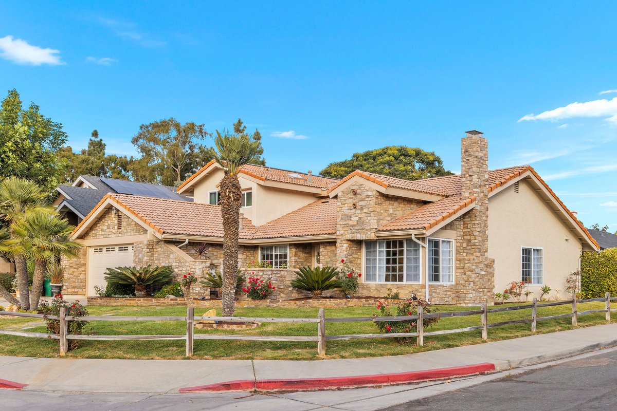 A house with a mix of stone and stucco exterior, landscaped yard with palm trees, bushes, and flowers, front sidewalk, and a street curb under a bright blue sky.