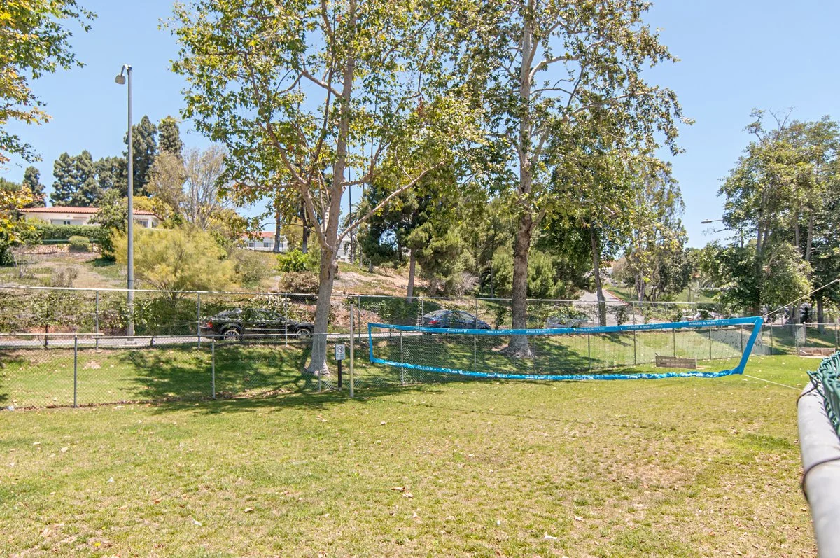 A fenced volleyball court with a net, surrounded by green grass and trees, with parked cars and houses in the background under a clear blue sky.
