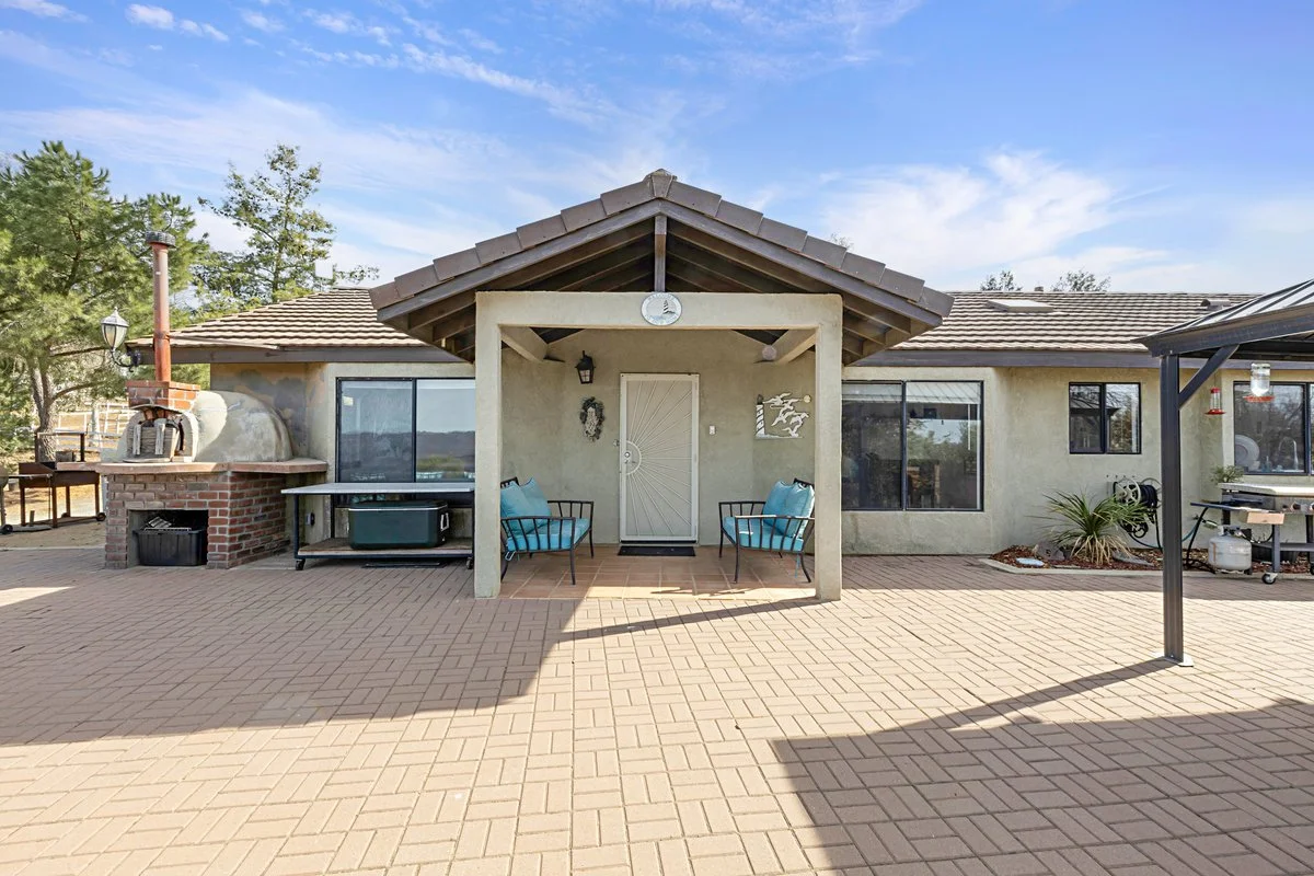Backyard patio with a covered entrance, outdoor seating with blue cushions, a brick pizza oven, a barbecue grill, and potted plants against a house with large windows and a blue sky in the background.