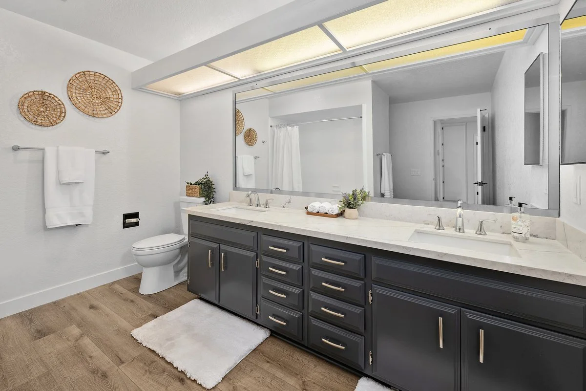 Modern bathroom with white walls, wood floor, large mirror, double sink vanity with dark gray cabinets, white marble countertop, and decorative baskets and plants. Towel bars with white towels, toilet, and shower area with curtain are visible.