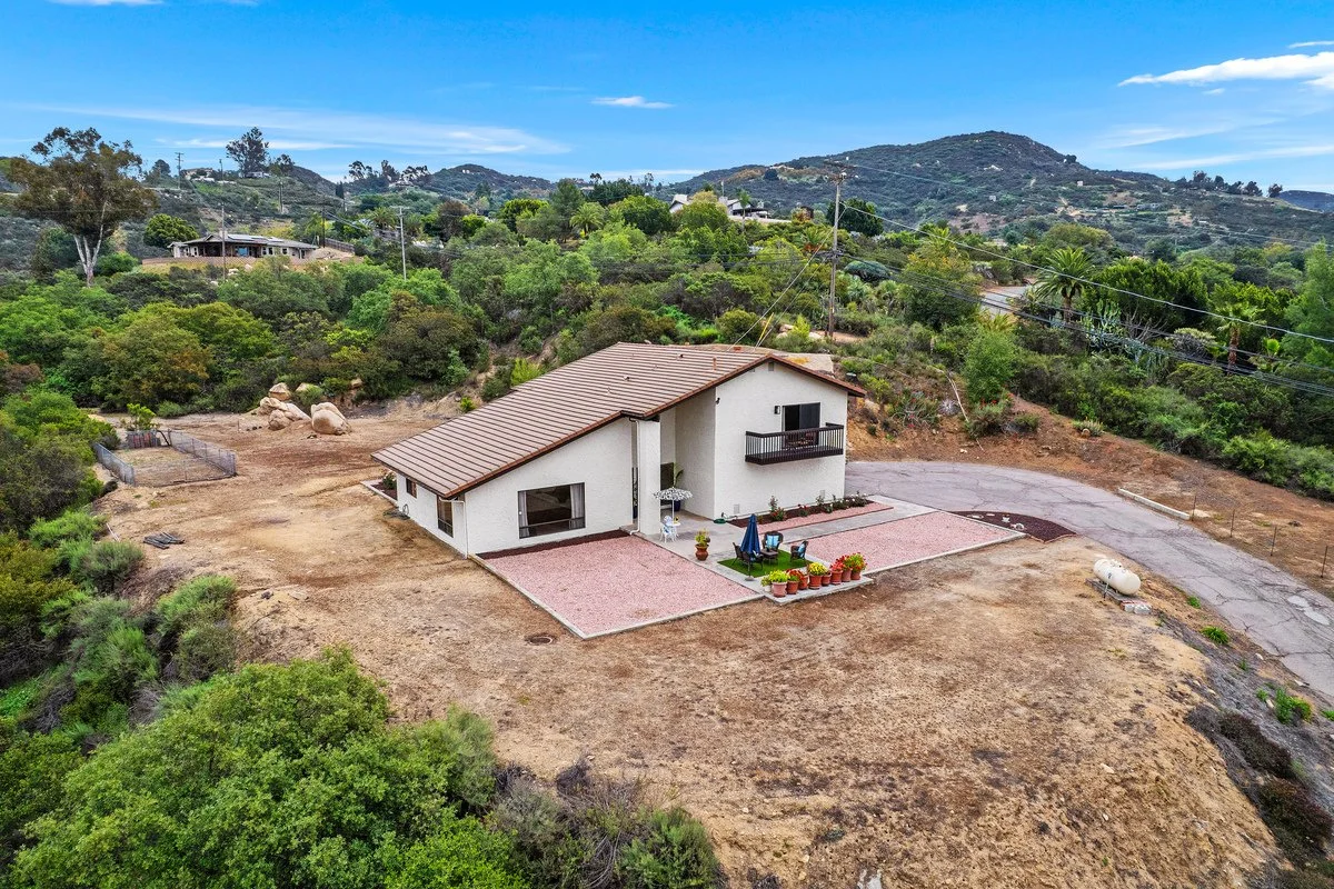 A white house with a brown roof located on a hillside with sparse grass and trees. There is a small patio with outdoor furniture and potted plants. Surrounding the house are hills covered with green vegetation and scattered houses.